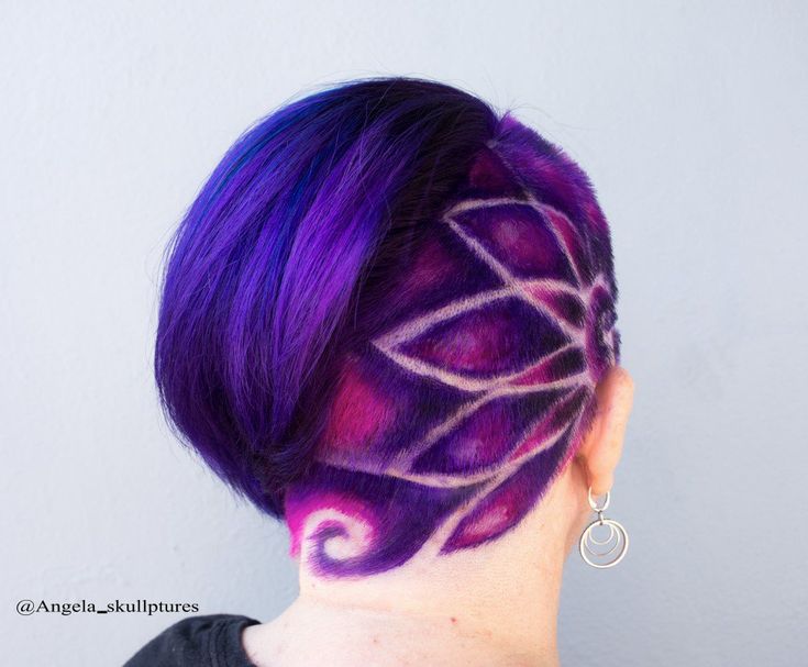 the image shows the back of a woman's head with a unique and artistic haircut. the hair is short, styled in a bob, and dyed in various shades of purple, ranging from dark bluepurple to lighter violet. on the shaved nape of the neck, there's an intricate floral pattern. the shaved portion is colored in purple and pink hues, with the design created by the contrasting skin color showing through. the woman is wearing a silver earring with a double hoop design on her right ear. the overall aesthetic is creative and bold, showcasing a personalized hair design.
