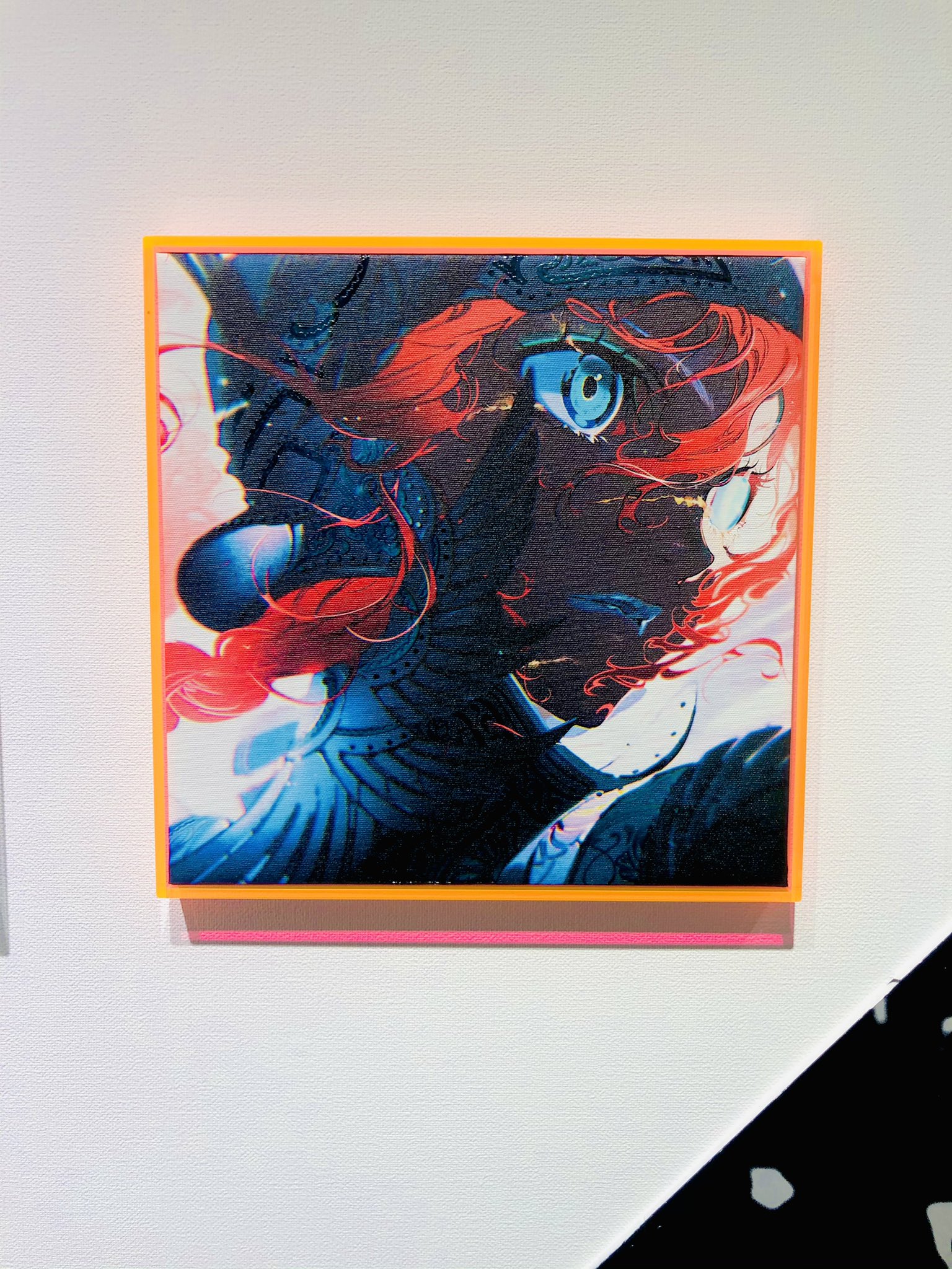 the image shows a framed piece of artwork. the art is a colorful, animestyle portrait of a person with red hair and a piercing blue eye. the portrait is set against a dark background with hints of white and blue, creating a sense of depth.  the art is framed in a bright orange frame that pops against the white wall. the lighting is artificial, likely from gallery spotlights, which highlights the vibrant colors and details of the artwork.