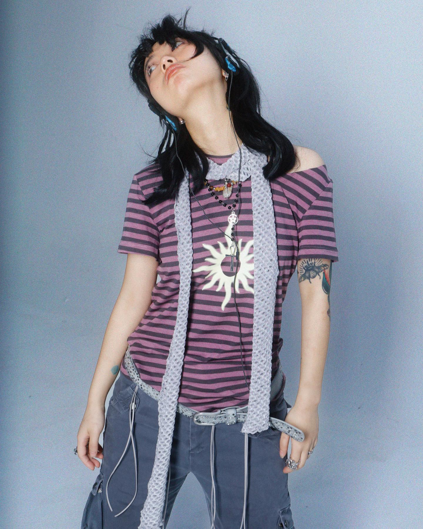a young asian woman with long black hair is shown from the waist up against a light grey background. she is wearing headphones, a purple and grey striped tshirt, grey cargo pants, and a long grey knitted scarf. she has a sunshaped pendant and other necklaces around her neck, and a small tattoo on her arm. her expression is slightly upward, as if she is listening to music and looking up. her style is casual but fashionable, with an alternative or indie vibe.