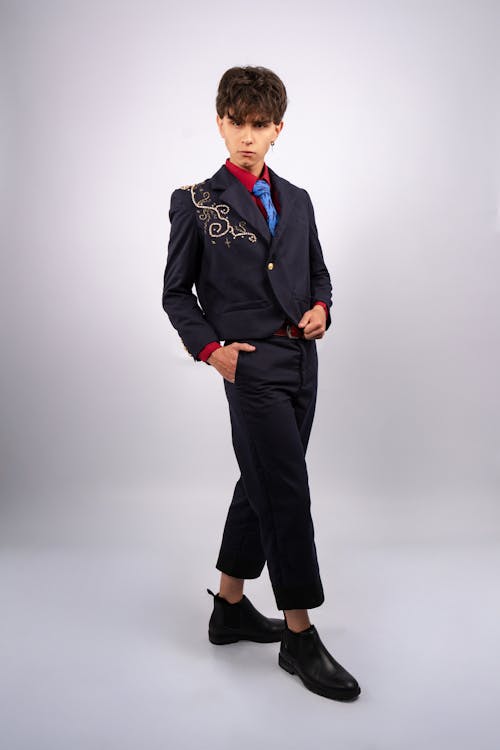 a man in a navy suit stands in front of a white background. the jacket has gold embroidery and is buttoned. he wears a red shirt and a blue tie. his hands are in his pockets. he wears black dress shoes.