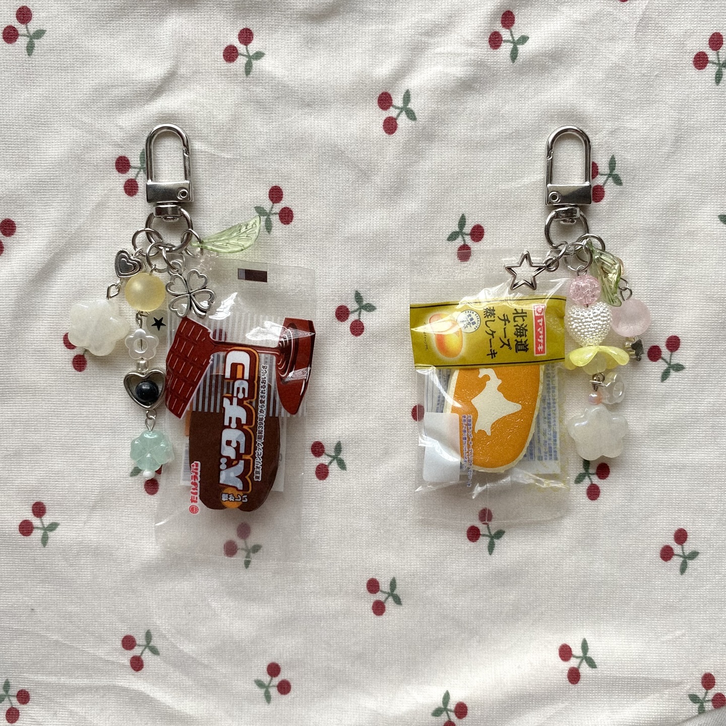 the image shows two keychains on a cherrypatterned fabric. each keychain features miniature, sealed snack or candy items along with decorative beads and charms. one keychain has a small package of "butter chocolate" and a green leaf charm, alongside beads and a clover charm. the other has a packaged slice of "peach cake" and a star charm, accompanied by decorative beads and a white cloud charm.