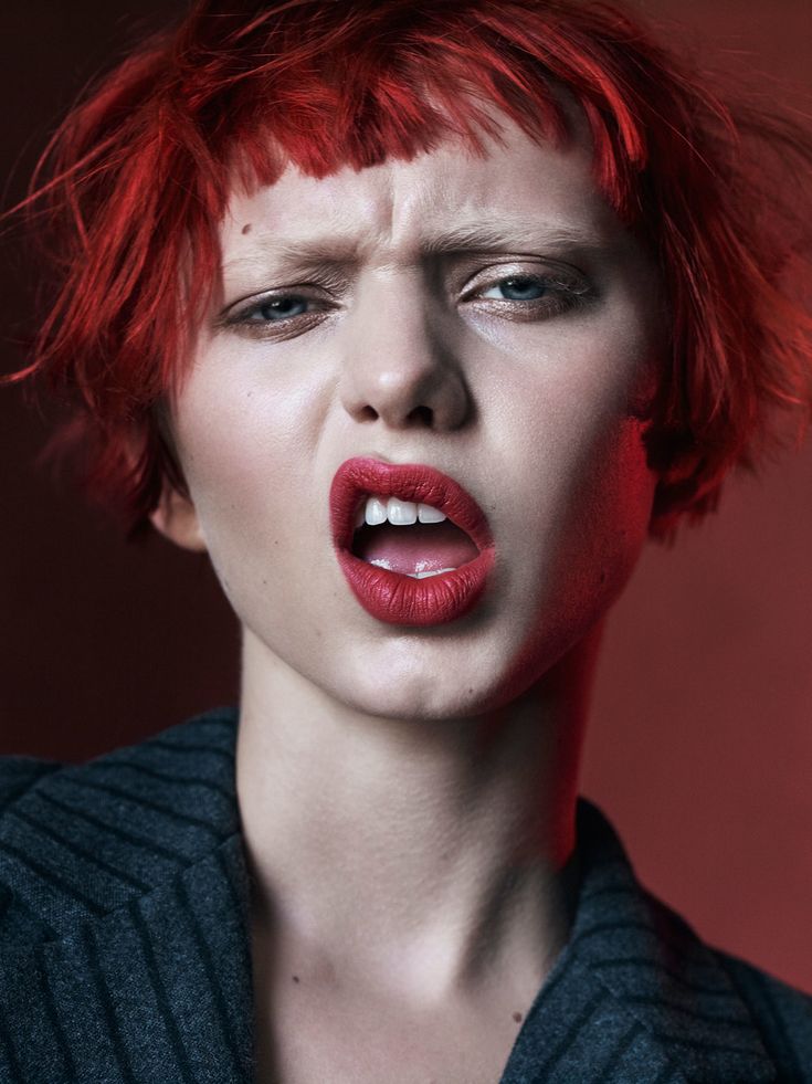 a closeup portrait of a female model with red hair. she has a short, choppy cut with a fringe. she is wearing red lipstick and has a serious expression on her face. she is looking directly at the camera. the background is a red and black gradient.  the photo is taken in a studio setting.