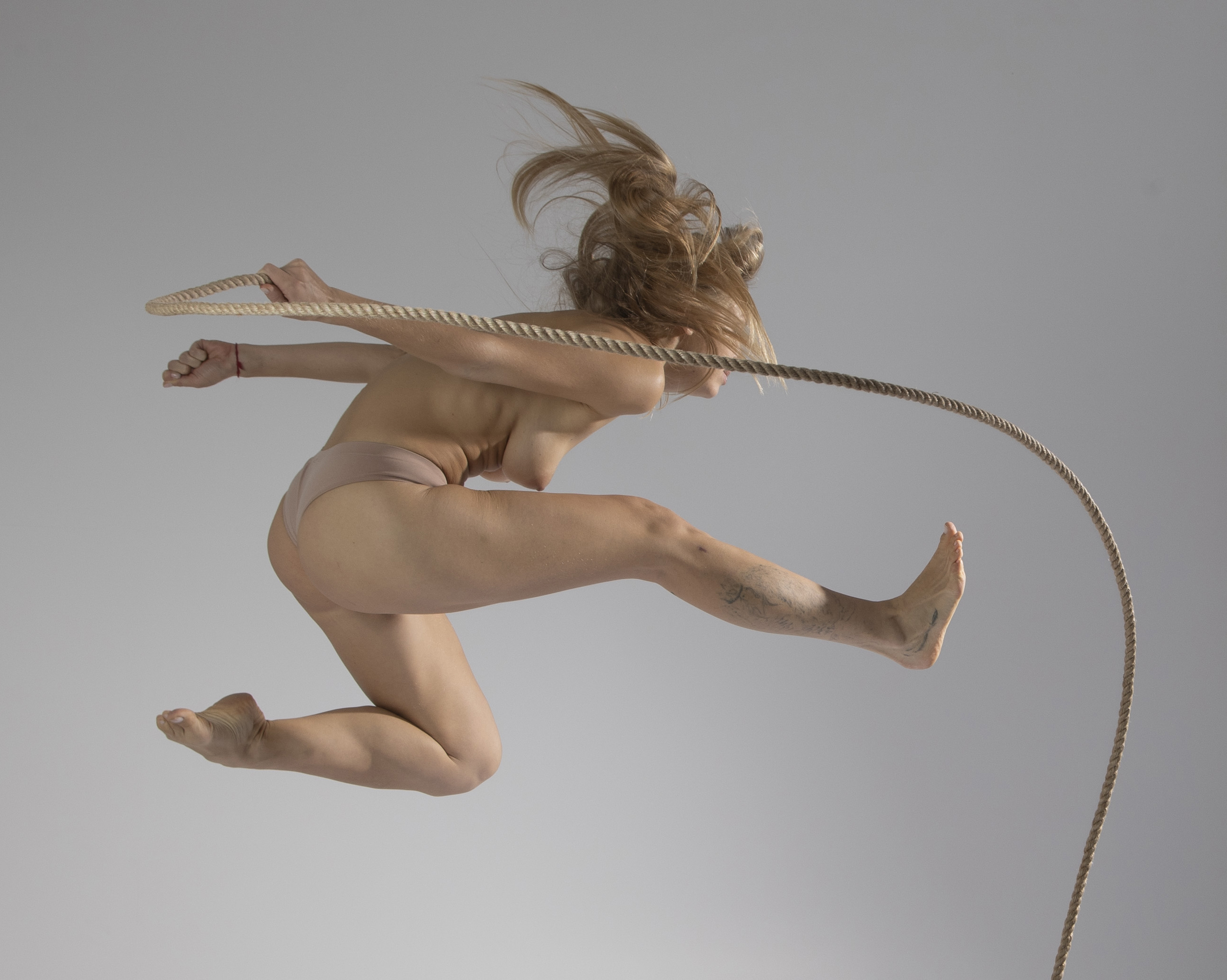 a full shot of a nude woman with blonde hair jumping and holding a rope against a gray background. she is barefoot and appears to be in motion. the image is taken in a studio setting.