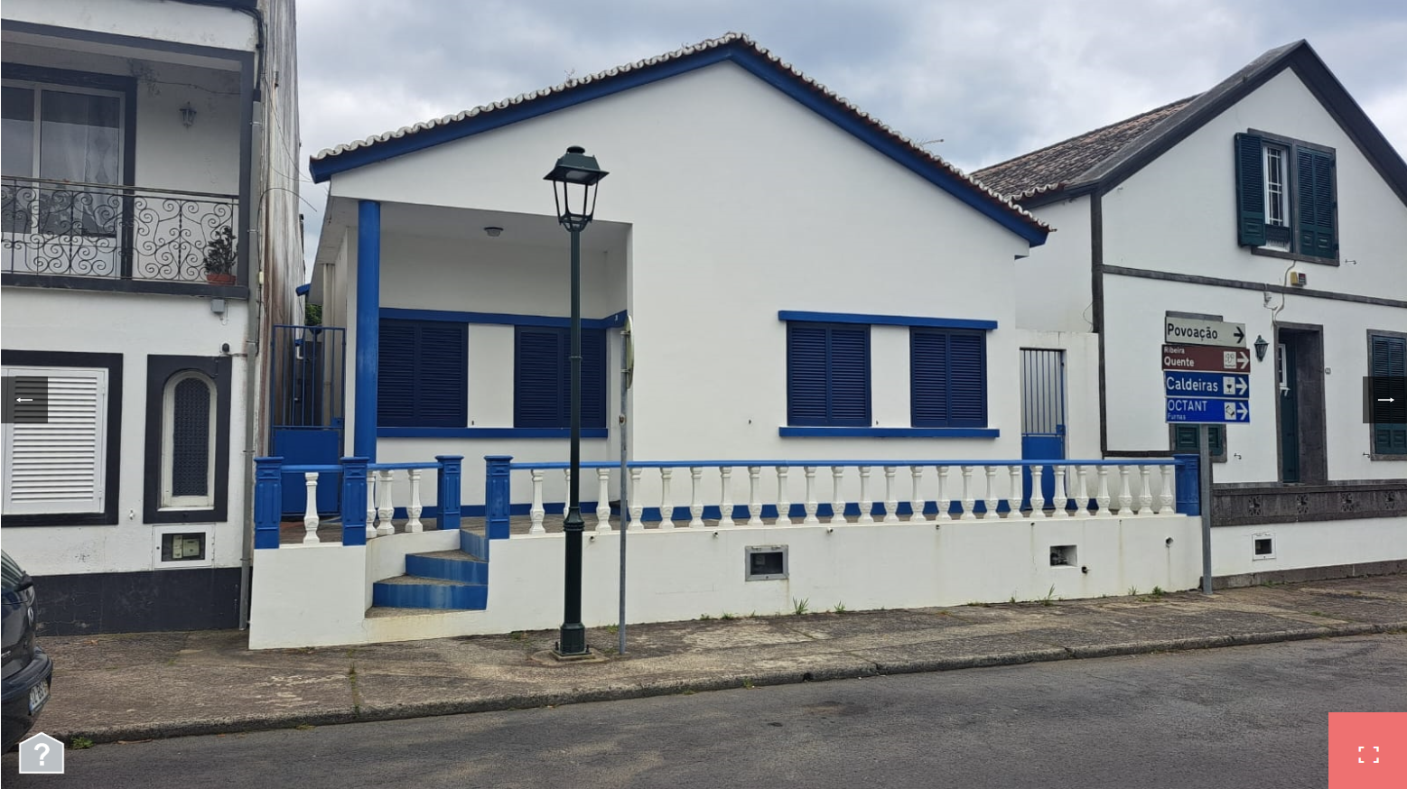 this image shows two white buildings with blue trim, one is a house and one is a commercial building with a street sign.  the house is on the left, and the commercial building is on the right. they are both in front of a street, with a sidewalk on the right side of the image, and a grassy area on the left side. in the foreground of the image is a sidewalk, and there is a lamp post in the middle of the street.  there are windows and doors on the exterior of the house, and a window on the exterior of the commercial building. there is a blue and white railing in front of the house.