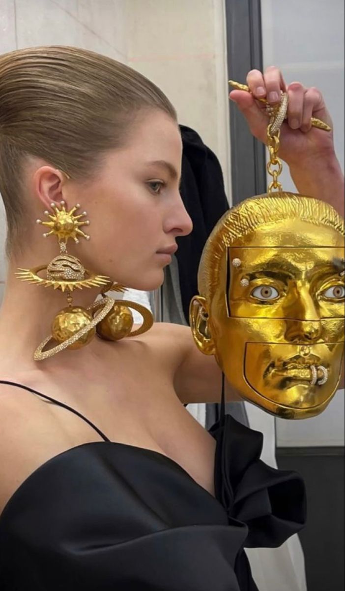 a woman with blonde hair styled in an updo is shown in a profile view, wearing large, elaborate earrings resembling planets. she is wearing a black dress. she is holding a golden purse shaped like a head. the purse has detailed facial features.