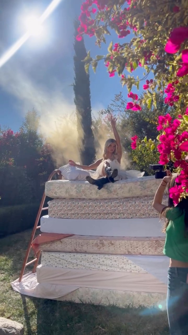 a woman in a white dress and boots sits atop a stack of mattresses in an outdoor setting, posing with one arm raised. a ladder leans against the mattresses. a photographer stands to the side, taking pictures. there are flowers and greenery in the background, and sunlight and smoke in the air.