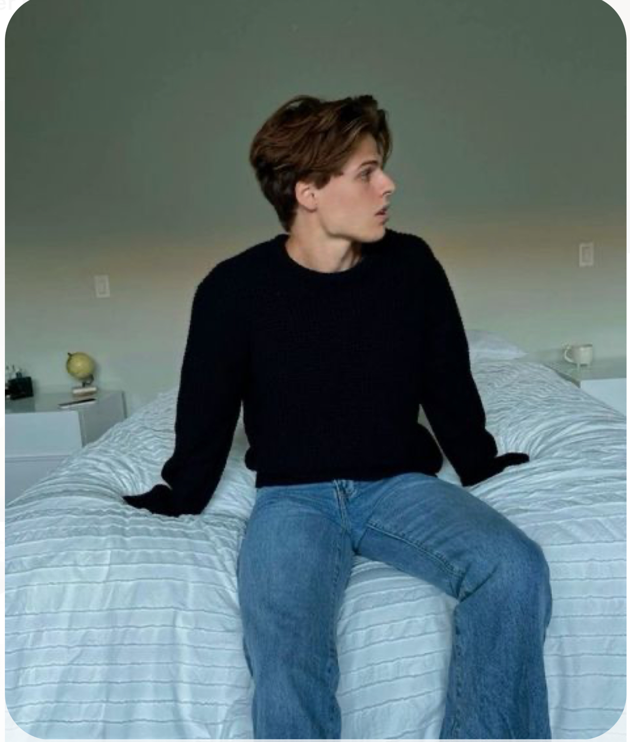 a young man with brown hair wearing a black sweater and blue jeans sits on a bed with white sheets. the walls are a muted teal, and there is a nightstand in the background. the image appears to be lit by natural light.