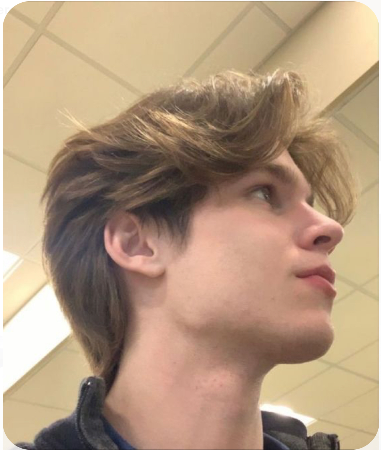 a closeup selfie of a young man with mediumlength, wavy blonde hair styled with a middle part. he has fair skin and is wearing a darkcolored shirt. the setting appears to be a public space with a drop ceiling and fluorescent lighting. the perspective is from a slightly low angle, capturing his side profile.