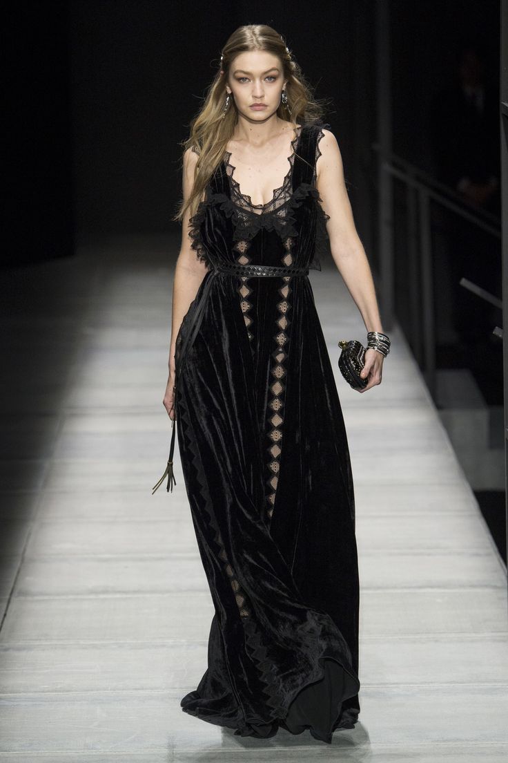 gigi hadid walks the runway in a stunning floorlength black velvet gown. the dress features intricate lace detailing and a belted waist. she accessorizes with a small black clutch and bracelets.  her hair is styled in loose waves, and her makeup is minimal but elegant, highlighting her features. the overall aesthetic is high fashion and glamorous, consistent with a fall/winter collection. the background suggests a professional runway setting.