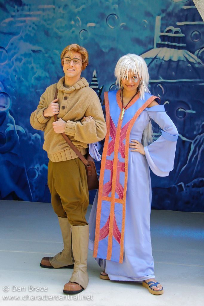 a full shot of a milo thatch and kida cosplay standing next to each other in costume with a blue background. milo thatch wears a beige, button up hoodie with rolled sleeves, brown shorts, brown boots, and brown eyeglasses. kida wears a light blue dress with an orange, red, and light blue applique down the front, sandals, and a necklace. milo has a brown shoulder bag over his shoulder, holding the strap. kida has her right hand on her hip.