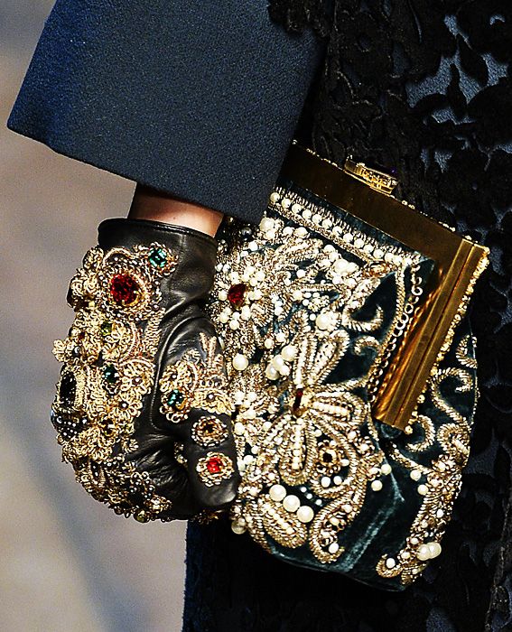 loseup shot of a woman's hand wearing an ornate, jeweled glove and holding a luxuriously embellished clutch purse. the glove and purse are heavily decorated with gold embroidery, pearls, and gemstones, creating a rich, opulent look. the background features a darkcolored, textured fabric. the overall image projects a sense of high fashion and luxury.