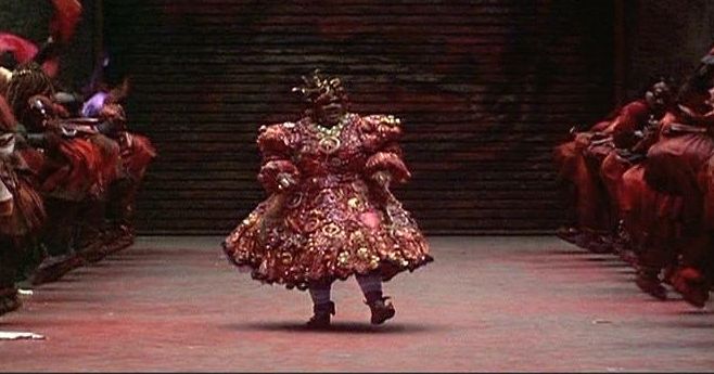 this is a movie still from the film "the wiz". the photo shows the character "the wiz" played by  actress diana ross, wearing a vibrant red costume with golden accents. the photo is taken from a low angle, focusing on the wiz standing at center stage. other figures in the background are also dressed in red and gold costumes, creating a sense of movement and energy, likely indicating a performance. the photo highlights the visual richness and theatrical elements of the movie.