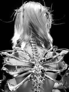 the image showcases a woman's back, adorned with an elaborate, metallic spinelike structure. this piece appears to be a fusion of fashion and art, resembling a sculpture. the lighting is studiobased, emphasizing the detail of the metallic piece against a stark black background.  the perspective is from the rear, focusing on the intricate design and how it interacts with the human form.