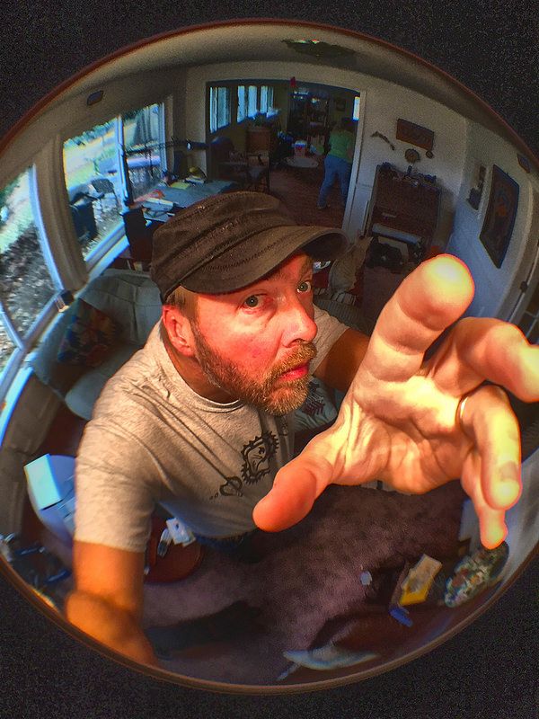 a man takes a selfie with a fisheye lens, showing his face, hand and part of his body. he is wearing a grey tshirt, jeans and a black baseball cap. he is smiling and has a goofy expression on his face. the image is distorted by the fisheye lens and the man's hand is stretched out in front of the camera.  the image is also taken from a low angle which makes the man's face look larger than it really is.  the background of the image shows the man's house, with a couch, table, and lamp visible. the wall is painted in a light color. the window is visible in the background.