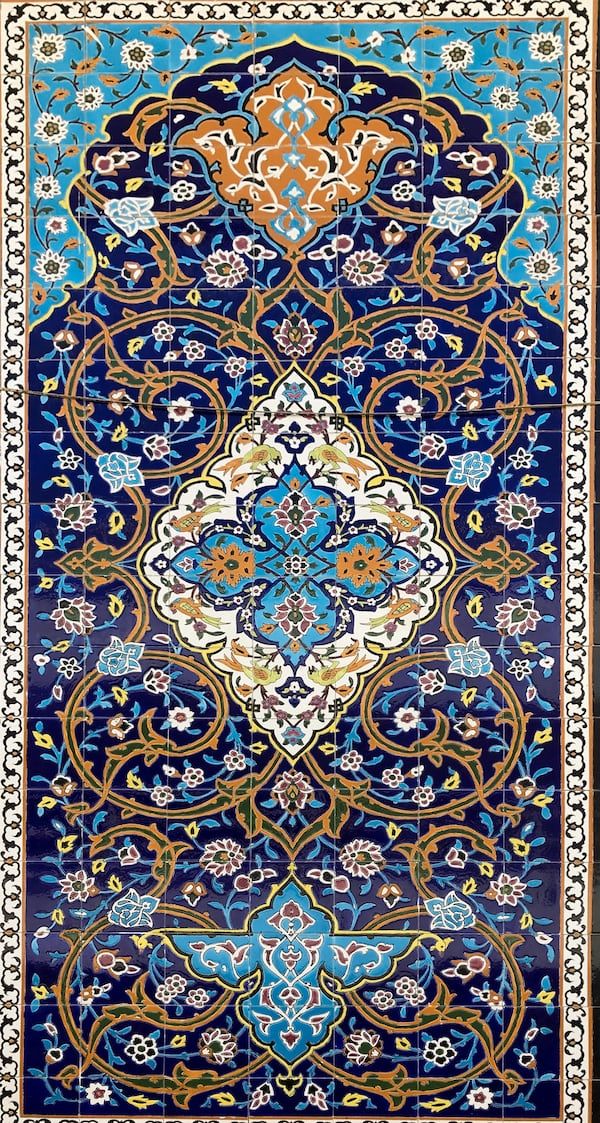 the image shows a panel of islamic mosaic tilework. the design features a repeating floral and geometric pattern in shades of blue, gold, white, and brown. the tiles are arranged to create a symmetrical composition with intricate details and a traditional persian art aesthetic. the artwork is framed by a border of smaller tiles.