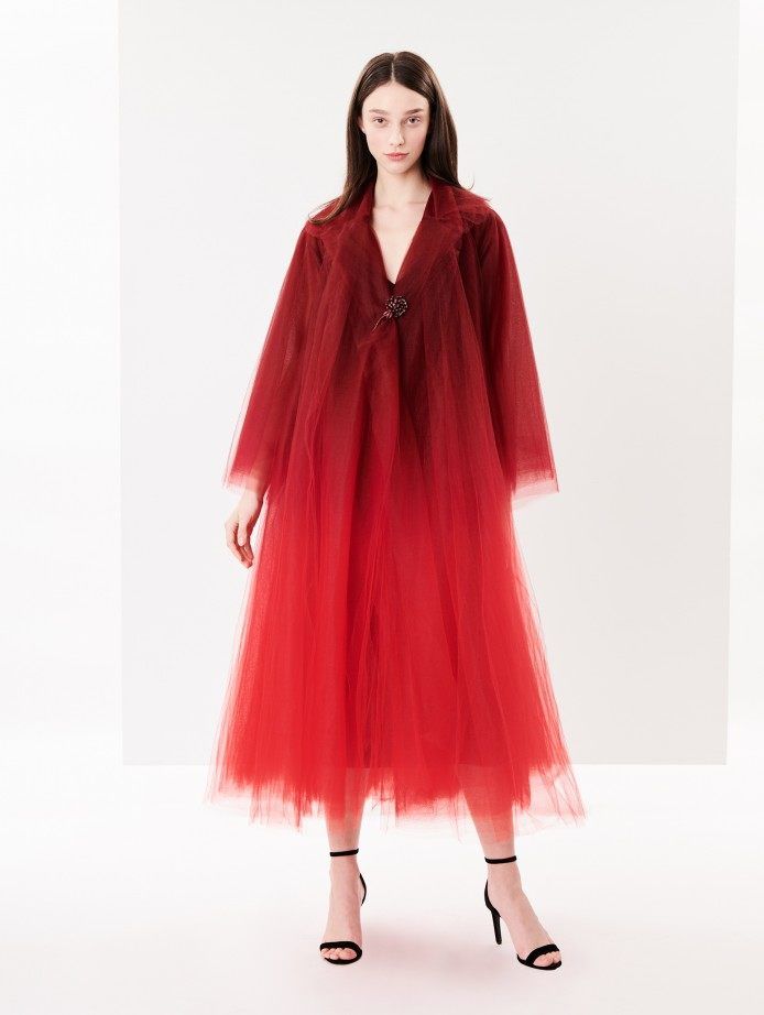 a woman with long brown hair is posing in a red ombre tulle dress with a vneckline and long sleeves. she's standing on a white background, her arms are relaxed and her face is looking straight at the camera. she's wearing black heels and a jeweled accessory on her dress.
