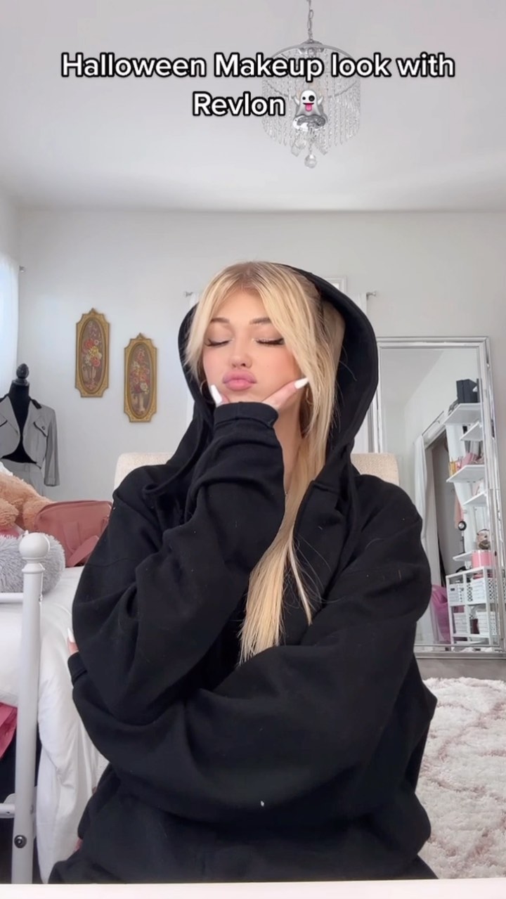 a young woman with blonde hair is posing in a black hoodie. the text "halloween makeup look with revlon" is at the top of the image. she is pouting and has white nail polish on. she is in a bedroom with a chandelier, bed, and other furniture.