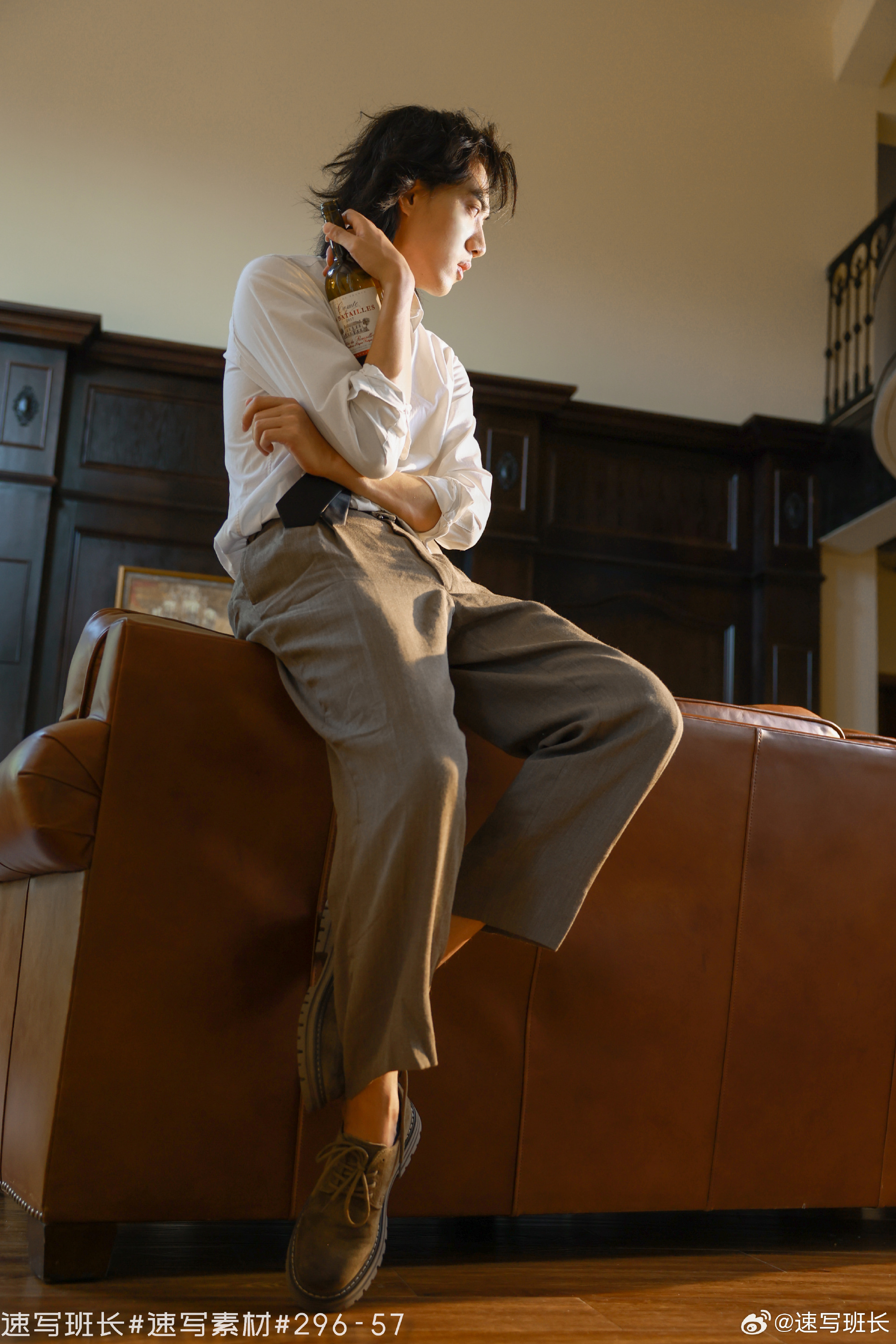 a young man with mediumlength black hair is perched on the back of a large leather couch, holding a bottle to his neck. he is dressed in a white buttondown shirt and loosefitting, highwaisted khaki pants. he is wearing brown leather shoes. the man is gazing downwards and to the right, offcamera. the room is luxurious, with a dark wooden wainscoting and a glimpse of a staircase in the background. the lighting is warm and inviting, coming from the windows on the right side of the frame.