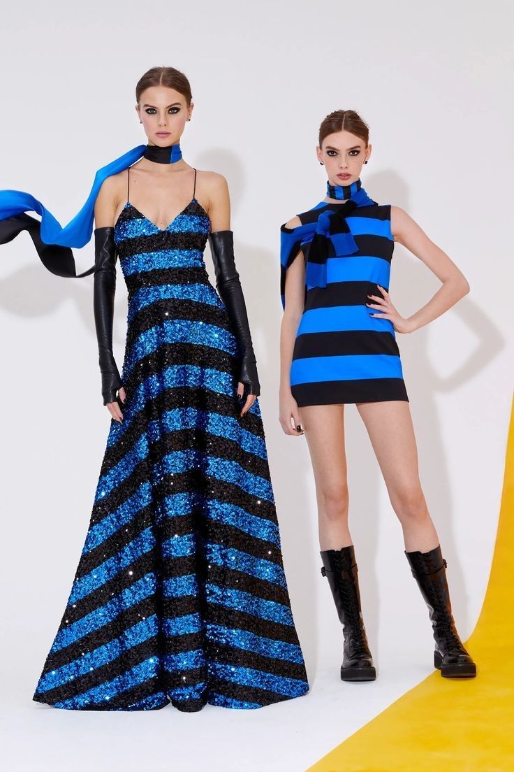 two women in blue and black striped dresses pose on a yellow and white runway. the woman on the left wears a sequined maxi dress and black gloves. the woman on the right wears a shorter dress and black boots. both women have a blue and black scarf around their neck.