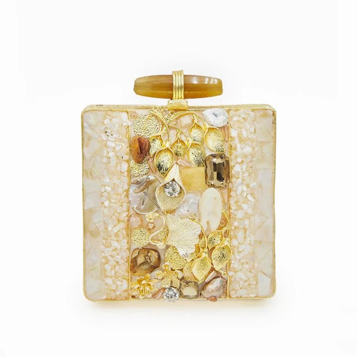 a square clutch handbag with a gold frame is adorned with a mosaic pattern along the sides and a central panel of decorative jewels, stones, gold leaves, crystals, and pearls. the top of the clutch features a light brown, stonelike handle with gold accents.