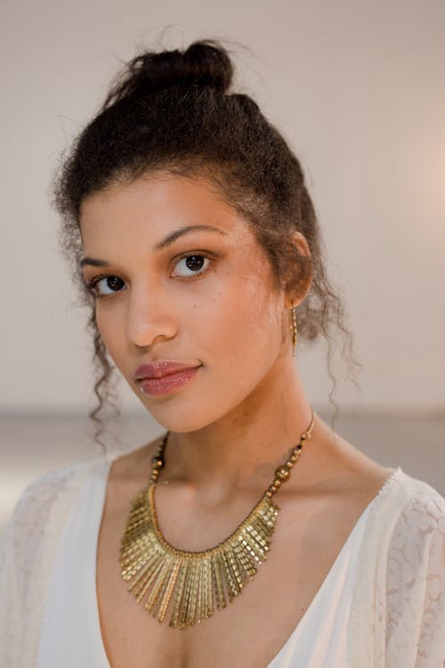 closeup portrait of a young woman with curly brown hair styled in a bun. she is wearing a white dress and a large, statement gold necklace.  her expression is serious and neutral. the lighting is soft and the background is blurred. the focus is on the woman's face and jewelry.