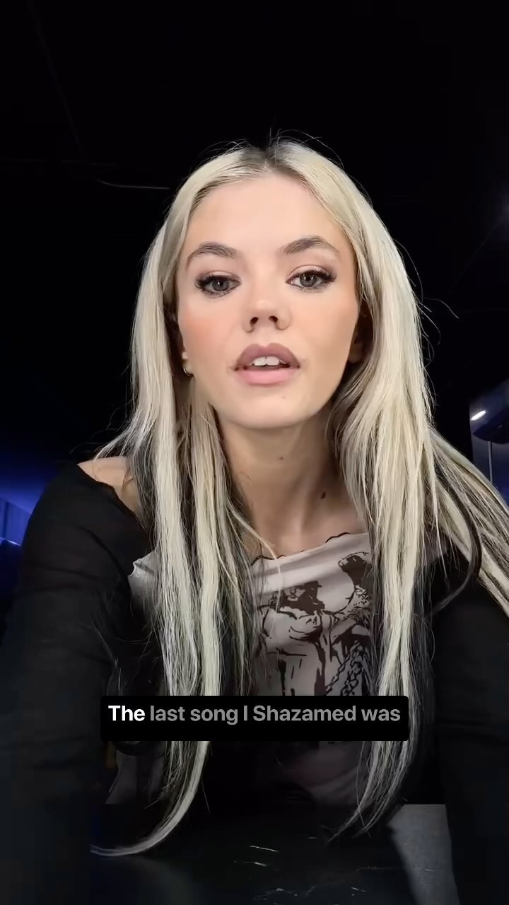 a woman with long blonde hair and black accents sits in front of a black background. the woman is wearing a longsleeved black shirt over a beige and black print shortsleeved shirt. she has fair skin and is looking into the camera and speaking. text on the bottom of the screen says "the last song i shazamed was."