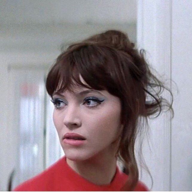 a closeup portrait of a woman with brown hair styled in a messy updo with bangs. she is wearing blue eyeliner and a red shirt, and her gaze is directed to the side.
