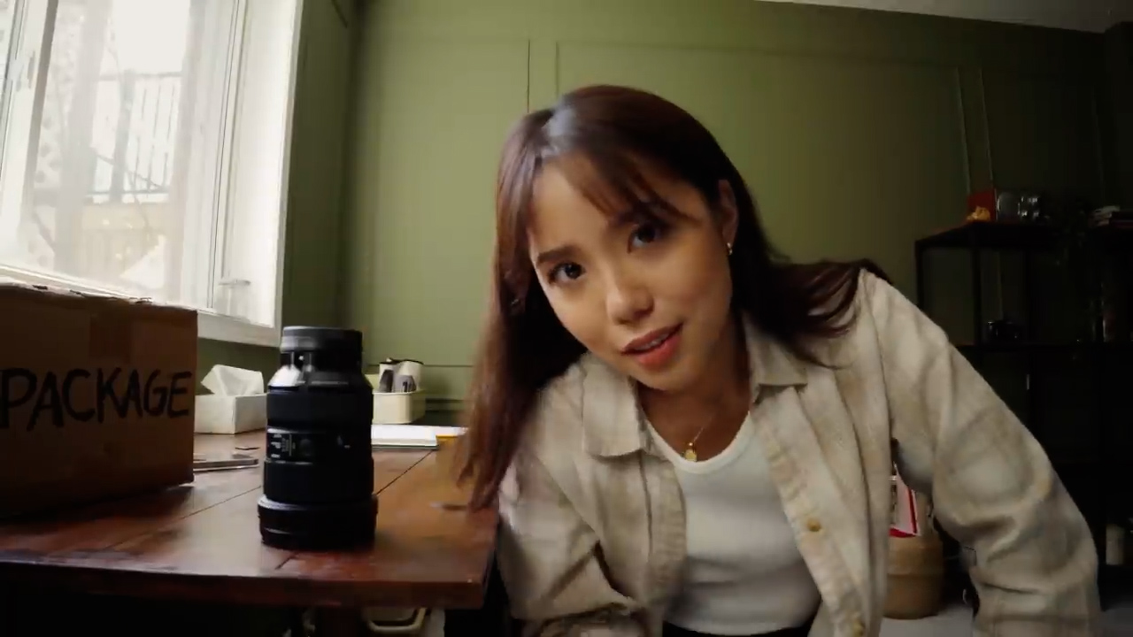 a young woman kneels beside a wooden table, engaging with the viewer. on the table sits a new camera lens, partially unboxed. the room is bathed in soft, natural light from a window out of frame. the overall atmosphere is relaxed and inviting.
