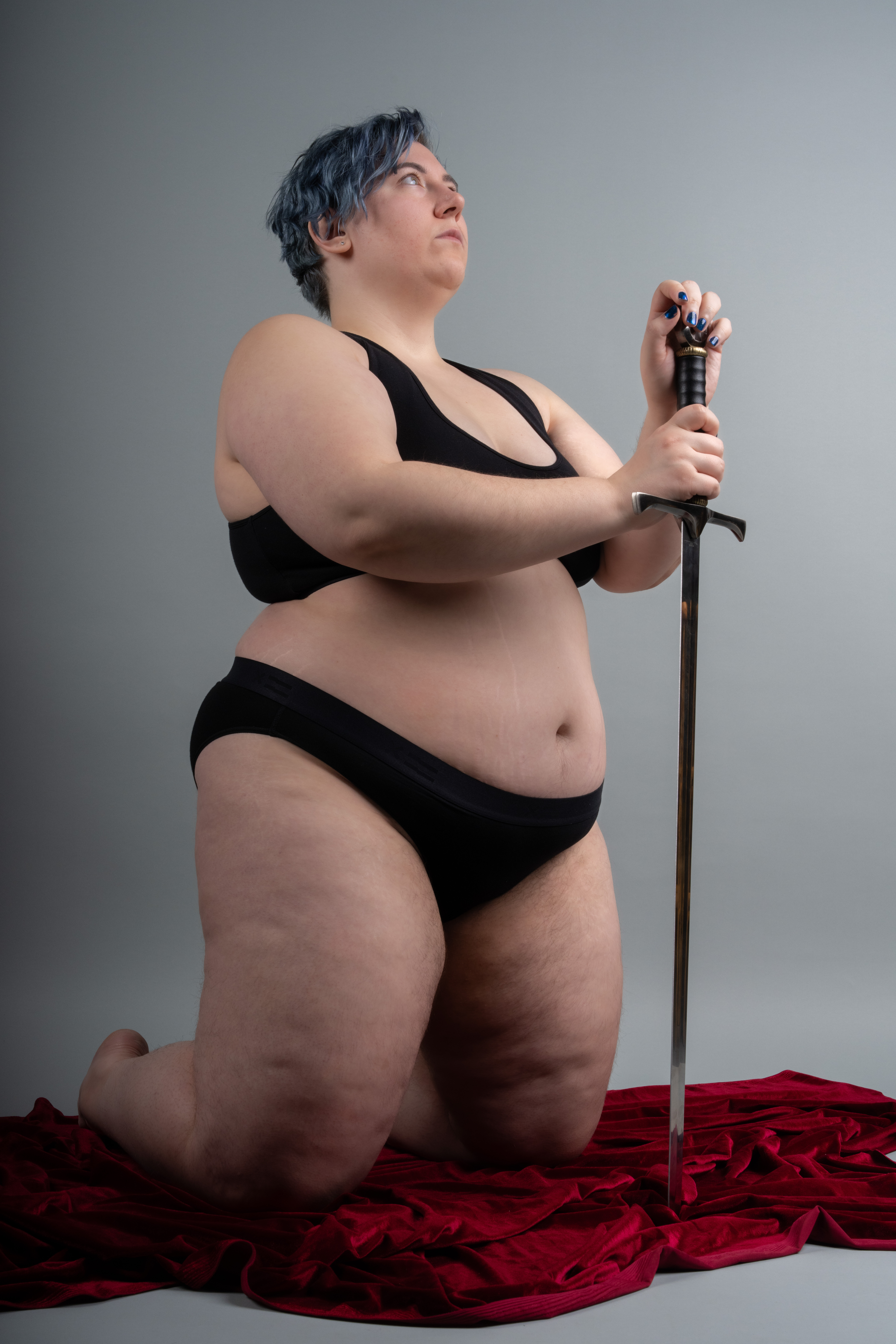 a plus size woman with blue hair is kneeling on a red velvet cloth, holding a sword. she is wearing a black bra and underwear. the background is a plain grey.
