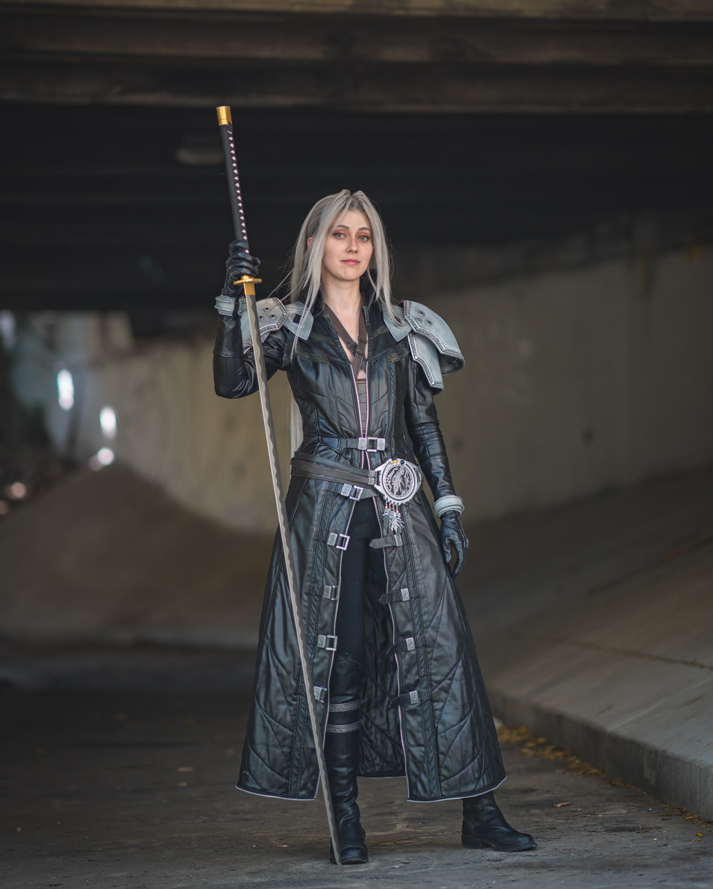 a striking image features a female cosplayer meticulously portraying cloud strife from final fantasy vii. the cosplayer is dressed in a detailed, accurate recreation of cloud's outfit, including the iconic long black leather coat adorned with silver buckles, shoulder pauldrons, and a matching belt with a detailed emblem. she holds a long katana, the weapon associated with cloud. the cosplayer's long, gray hair and stoic expression further enhance the portrayal of cloud's character. the setting is a dark urban underpass, which adds to the moody atmosphere of the photograph. the overall aesthetic is wellexecuted, capturing the essence of the character and the game's visual style.