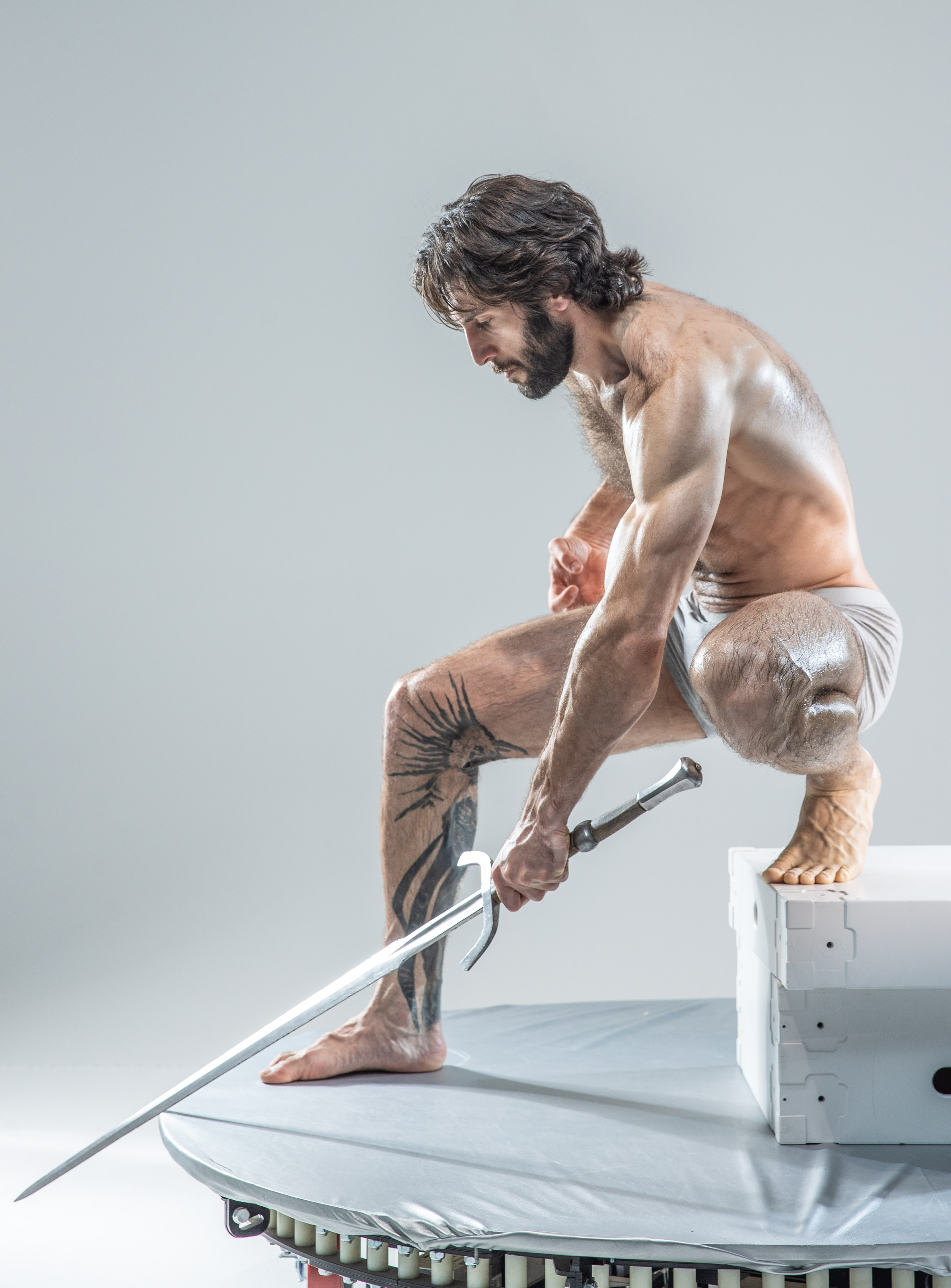 a muscular man with long dark hair and beard is posed dynamically on a white platform. he is holding a long sword in his right hand, the blade extending towards the left of the image. his left hand is clenched in a fist. he is lit by studio lighting which casts soft shadows and highlights his physique. the background is a plain gray.