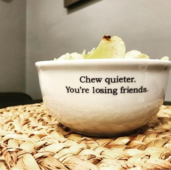 a white bowl filled with potato chips sits on a woven mat. the bowl has black text that reads, "chew quieter. you're losing friends." a few chips are sticking out above the rim of the bowl.