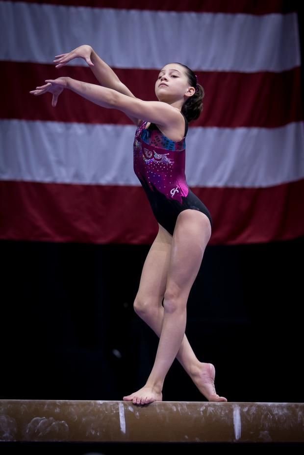a young female gymnast performs a routine on the balance beam during a competition. the american flag is visible in the background. the gymnast is wearing a black leotard with pink and purple accents. she is focused on her movements and her athleticism is evident.