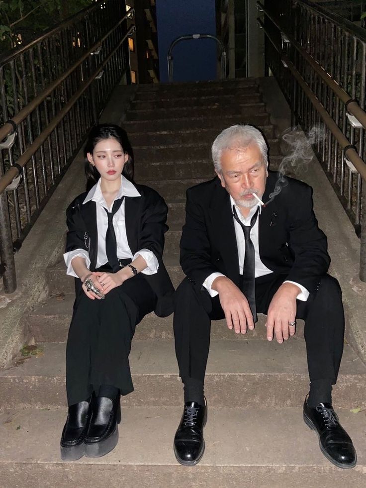 a young woman and an older man, both dressed in black suits and ties, sit on the stairs of an urban setting at night. the woman is wearing a white shirt and has her hair pulled back. the man has a white shirt, tie, and is smoking a cigarette. they are both looking at the camera. the image has a grunge aesthetic, with the stairs and surrounding environment looking worn and weathered. the couple exudes a cool, edgy, and stylish vibe.