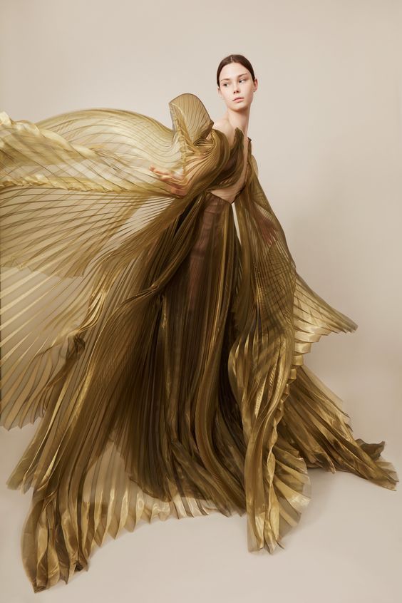 a woman in a gold pleated dress poses elegantly in a studio setting. the dress is flowing and ethereal, creating a striking and beautiful image.