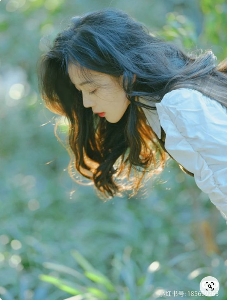 he image captures a young woman with long, dark hair, wearing a white blouse. she is outdoors, amidst lush green foliage, her head bowed as if in contemplation. the sun shines through the trees, creating a soft, dappled light that illuminates her hair and the surrounding greenery. the overall ambiance is serene and peaceful.