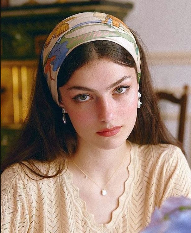 the image is a closeup portrait of a young woman with long, brown hair styled with a vintageinspired floral patterned silk scarf as a headband.  she's wearing pearl earrings and a delicate gold pearl necklace. her clothing consists of a light beige, knit sweater with a scalloped neckline. she has a calm and serene expression. the overall aesthetic is soft, elegant, and reminiscent of vintage or retro styles. the background is blurred but shows a hint of an indoor setting with warm, natural lighting.