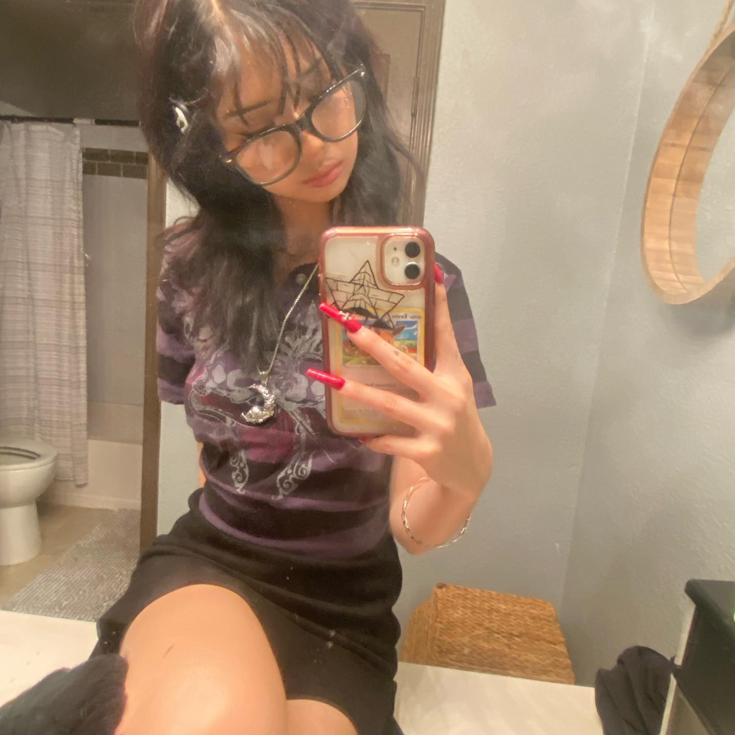 a young woman with dark hair and glasses poses for a mirror selfie in a bathroom. she is wearing a purple and black graphic tee, black skirt, and silver jewelry. she has red nails and is holding her phone in front of her face. the bathroom is welllit and has a neutral color scheme.