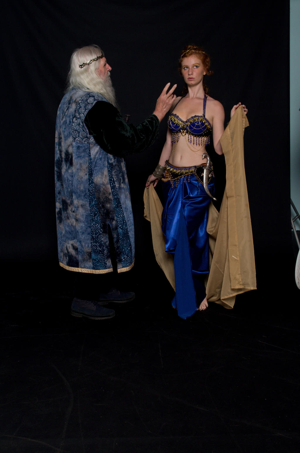 a full shot of a woman dressed as a belly dancer in a blue and gold costume holding a gold sheer fabric while standing next to a man dressed as a wizard in a blue and black gown and headdress against a black background.