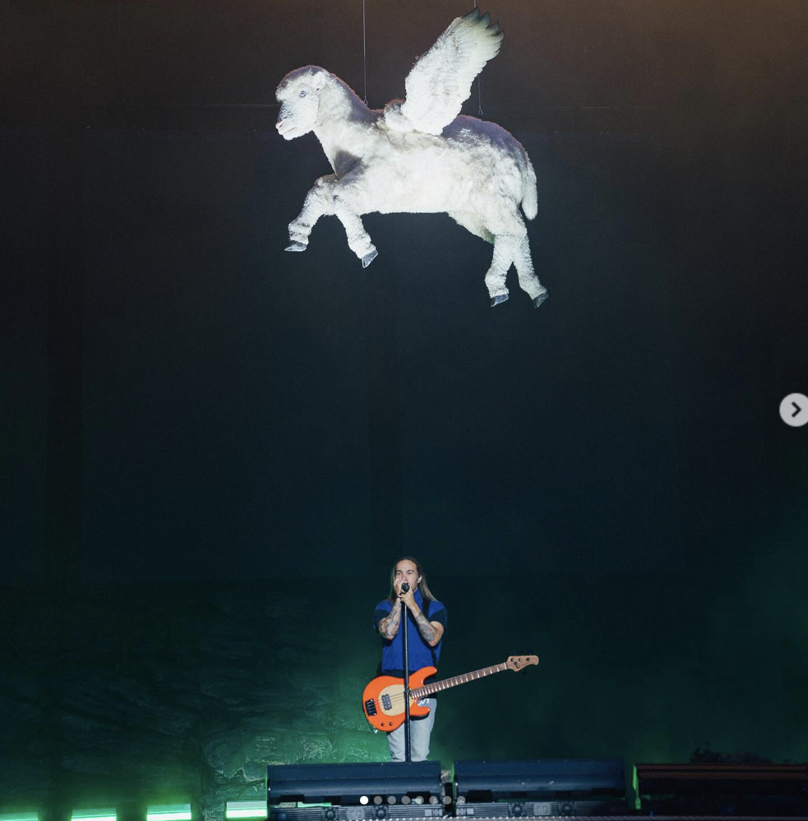 a musician with a bass guitar is performing on stage in front of a large crowd, lit by a bright spotlight. the performer is singing into a microphone while playing their instrument. in the background, a large, white, flying sheep hangs from the ceiling.