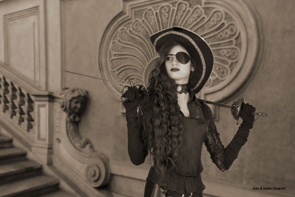 a black and white image of a woman in a piratestyle costume. she is wearing an eyepatch, a large hat, a corset, lace gloves, and a choker. she is holding two swords, a rapier in each hand. she is standing in front of a staircase in an antique or baroque building. the image has a vintage, historical feel.