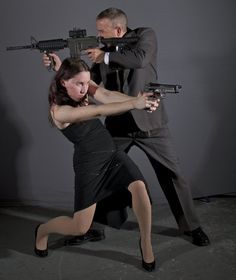 a man and a woman are posing with guns in what appears to be a studio setting. the man is standing behind the woman, holding a rifle. the woman is in front, holding a handgun. both are aiming their weapons forward. the man is wearing a suit, while the woman is wearing a black dress and high heels. the background is a plain gray. they could be posing for a promotional image for a movie or a play.