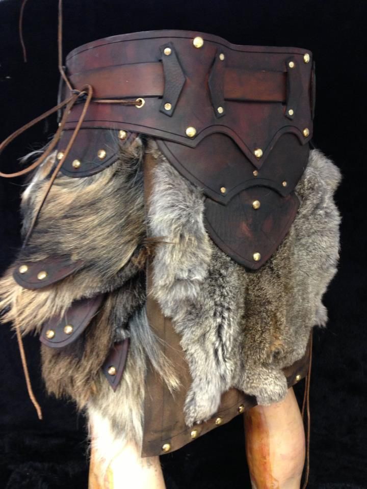 the image shows a piece of handcrafted medievalstyle armor. it features a wide, brown leather belt adorned with geometric leather appliques and goldcolored rivets. hanging below the belt are sections of fur, arranged in layers, interspersed with smaller leather pieces also studded with rivets. below the fur, a leather skirt extends down to the legs, which appear to be those of a mannequin. the armor evokes a rustic, historical feel reminiscent of viking or renaissance attire.