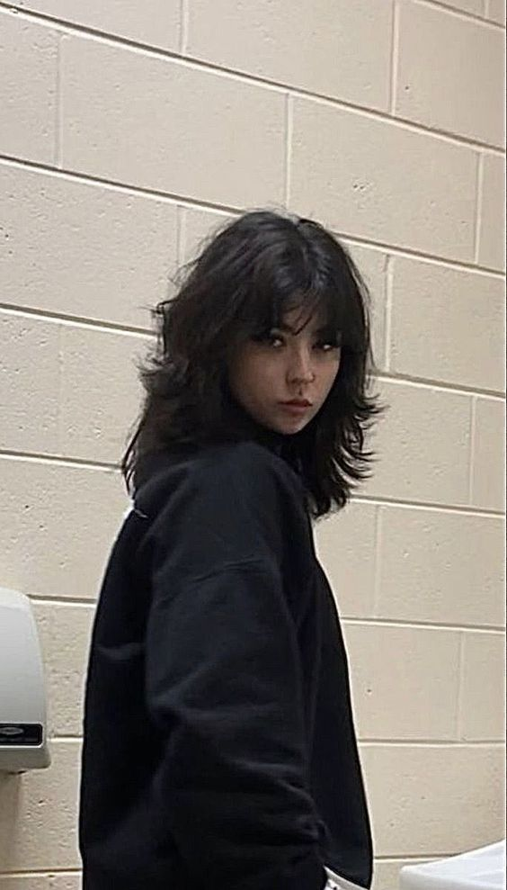 a young girl with black hair is standing in a bathroom against a white tiled wall. she is wearing a black sweatshirt and is looking at the camera with a serious expression.