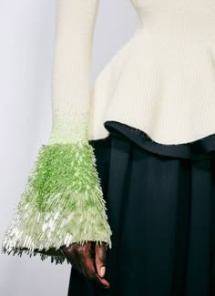 a woman is wearing a white sweater with a black skirt. the sweater has long sleeves with green fringe detailing. the woman is looking to the left, while her hand is on her hip. the image is taken from a slightly low angle, which emphasizes the length of the skirt and the dramatic nature of the sweater's sleeves.