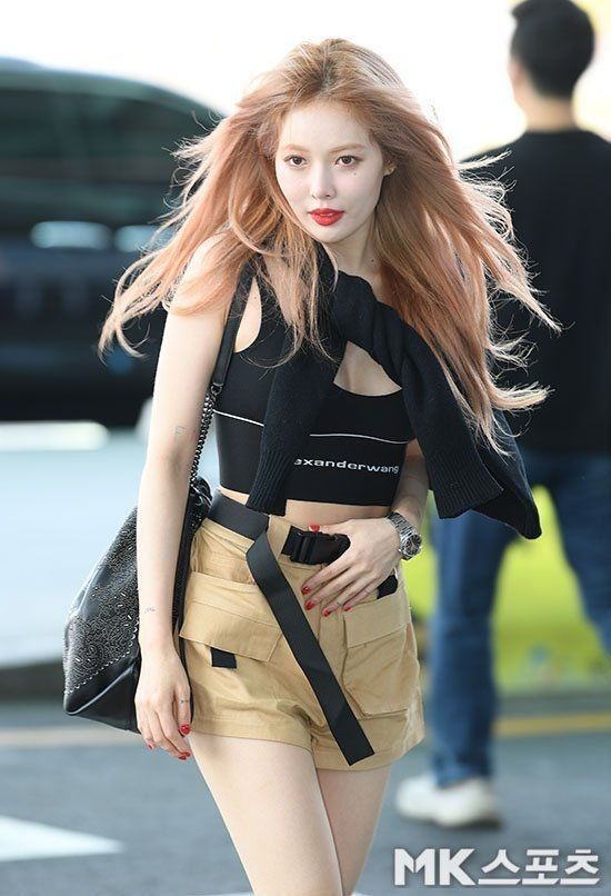 a young woman is walking through an airport, wearing a stylish outfit that includes a black crop top, khaki shorts, and a black leather bag.  her long blonde hair is flowing in the wind and she is wearing a small pair of sunglasses. she has a red lipstick.