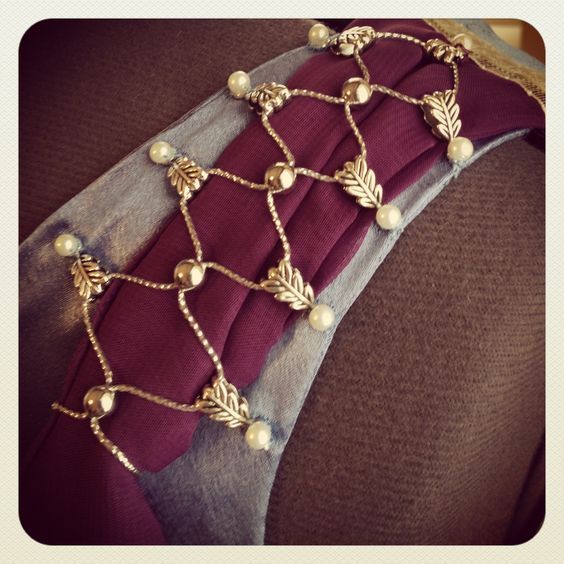 a closeup shot displays a necklace laid upon layered fabric. the necklace features a pattern of gold leaves and pearls connected by thin gold wire forming geometric shapes. the underlying fabrics consist of a maroon, pleated layer centered between two light blue layers on each side. the entire composition rests on a brown, textured surface.