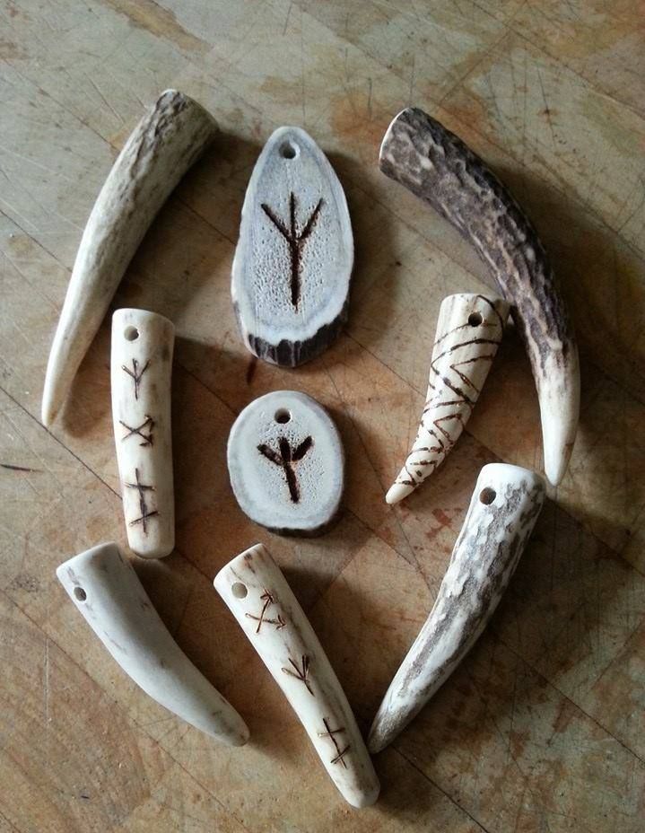 the image shows an arrangement of rune stones and pendants made from antler and horn on a wooden surface. some pieces are carved with runes or other symbols, and some are natural antler pieces with drilled holes for use as pendants.