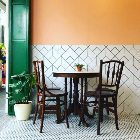 a cafe interior with two chairs and a table with a potted plant on top. the walls are orange and white tiled, with green on the left. there are two chairs and a table. the chairs are wooden and have a black finish. the table is made of wood and has a white top. the walls are tiled with white tiles. there are two potted plants in the image. the floor is tiled with white tiles.