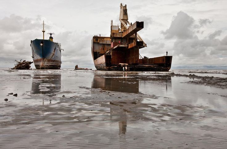 the image depicts a scene at a ship breaking yard, likely in chittagong, bangladesh.  two large ships are prominently featured. one is a relatively intact, though aged, vessel grounded on the mudflats. the other is a significantly dismantled and rusting ship, partially submerged, with workers visible amidst the wreckage. the beach is largely exposed due to low tide, revealing muddy textures. the reflection of the ships and sky is visible in the shallow water. the overall atmosphere is somber and gray, reflecting the harsh environmental conditions and the nature of the shipbreaking process.